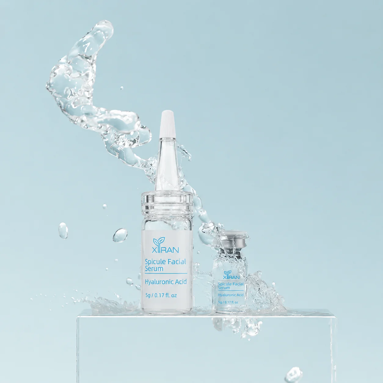Two bottles of Xiran Spicule Facial Serum displayed on a clear block with water splash in a clean light blue background.